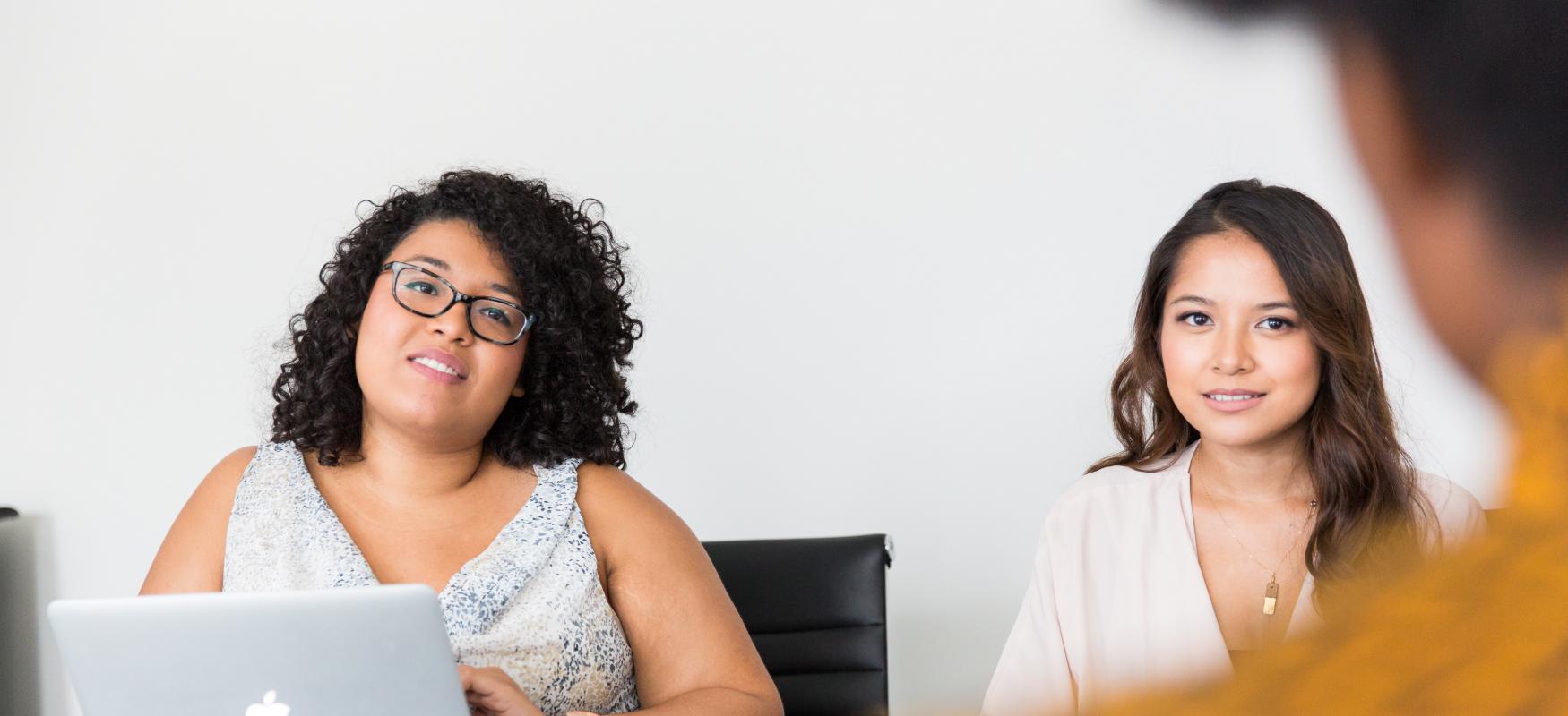 three women sitting around a conference table having a discussion, one with a computer on the table in front of her.
