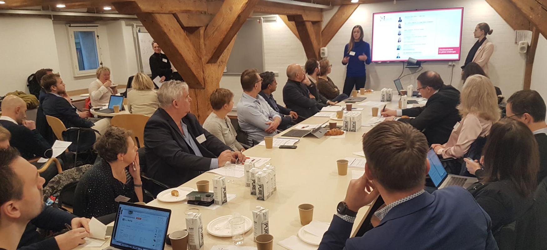 Participants gathered around a large table at a workshop and a person speaking in front of them.